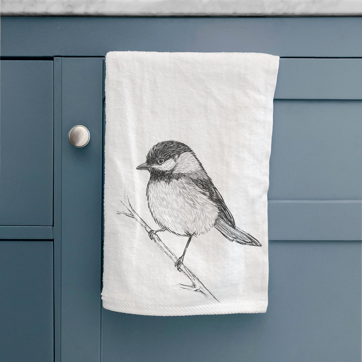 White hand towel printed with a detailed black ink sketch of a black-capped chickadee perched on a branch, hanging on a cabinet door.