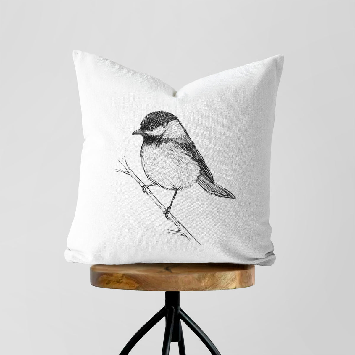 White pillow cover printed with a detailed black ink sketch of a black-capped chickadee perched on a branch, on a wooden stool.