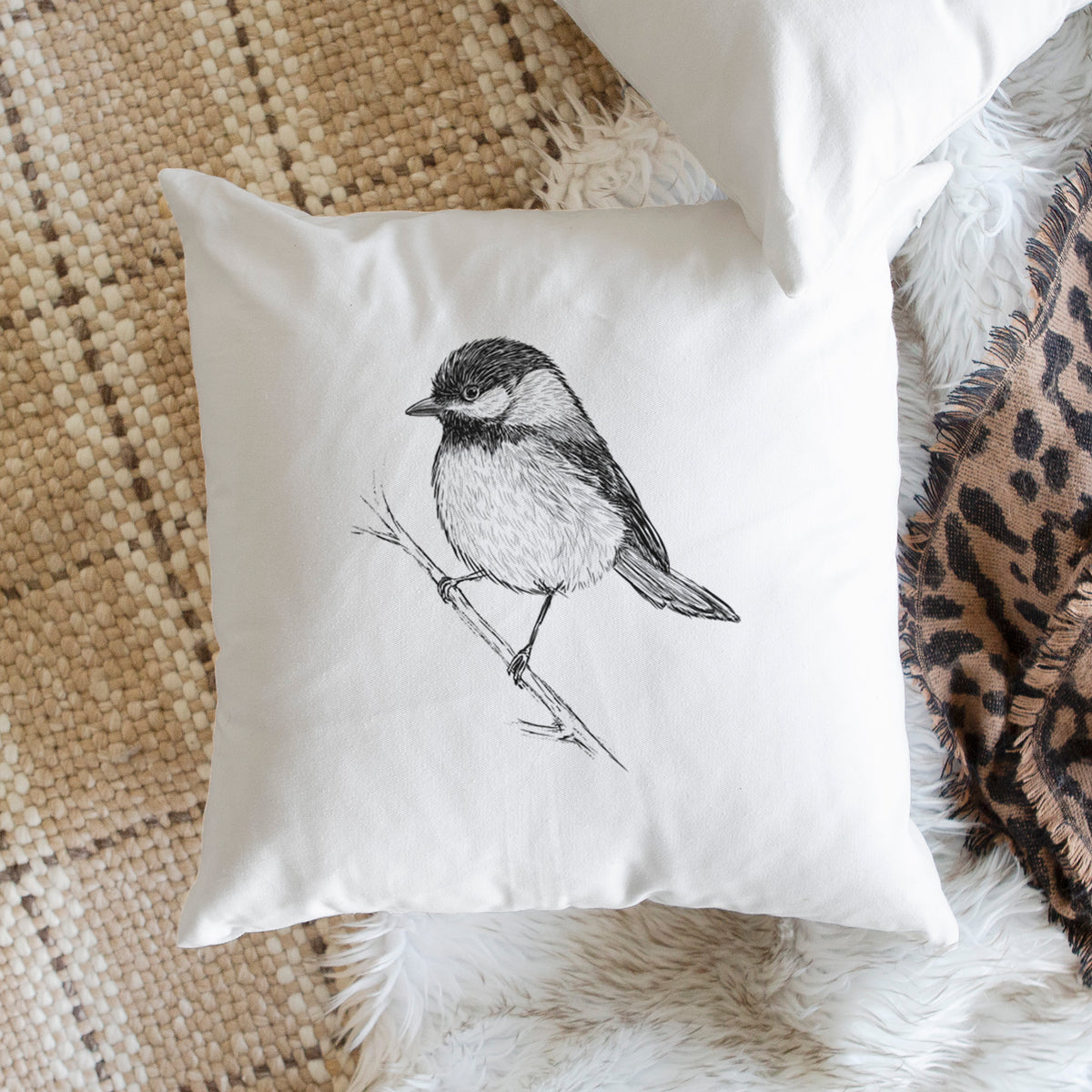 White pillow cover printed with a detailed black ink sketch of a black-capped chickadee perched on a branch, on a woven chair.