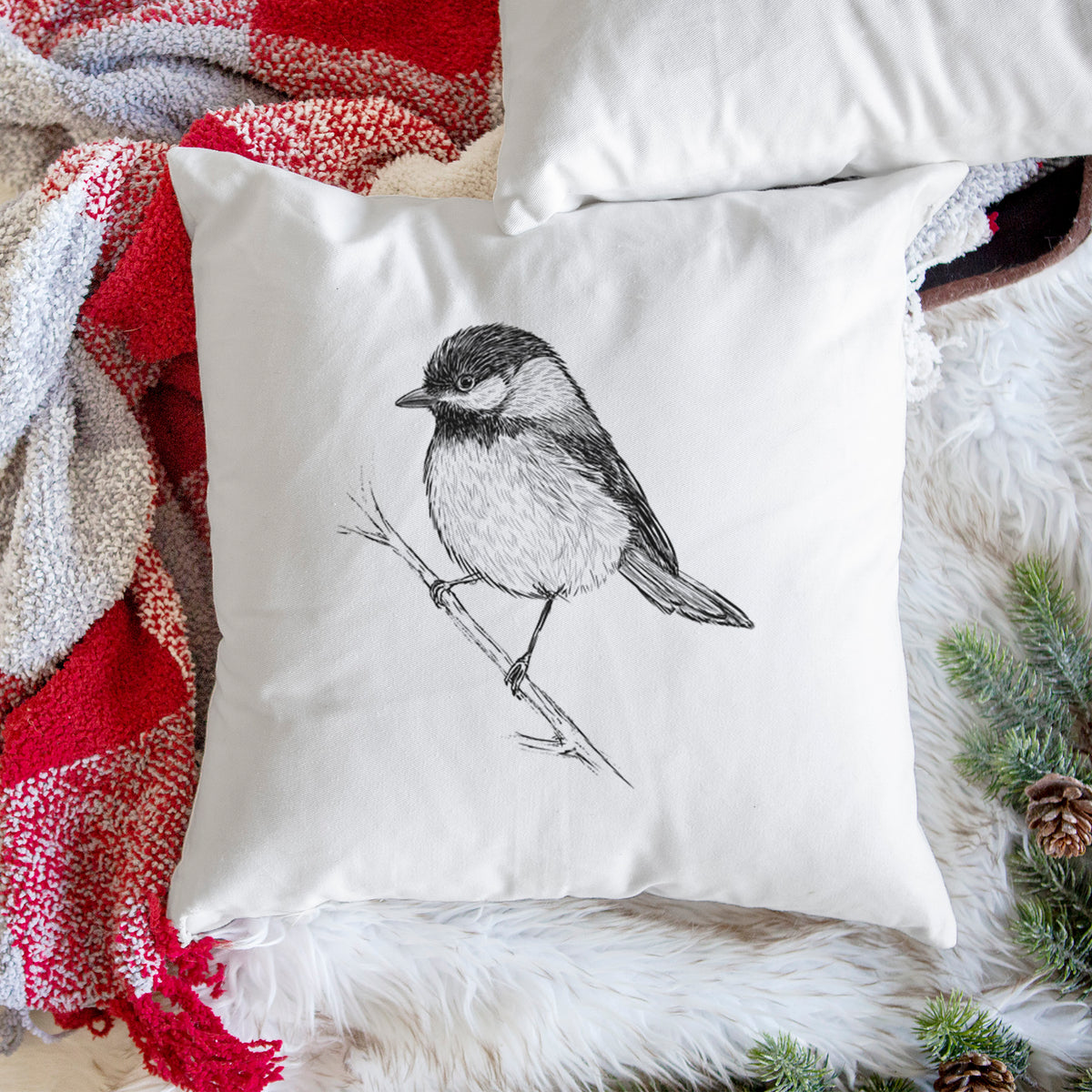 White pillow cover printed with a detailed black ink sketch of a black-capped chickadee perched on a branch, on a woven chair.