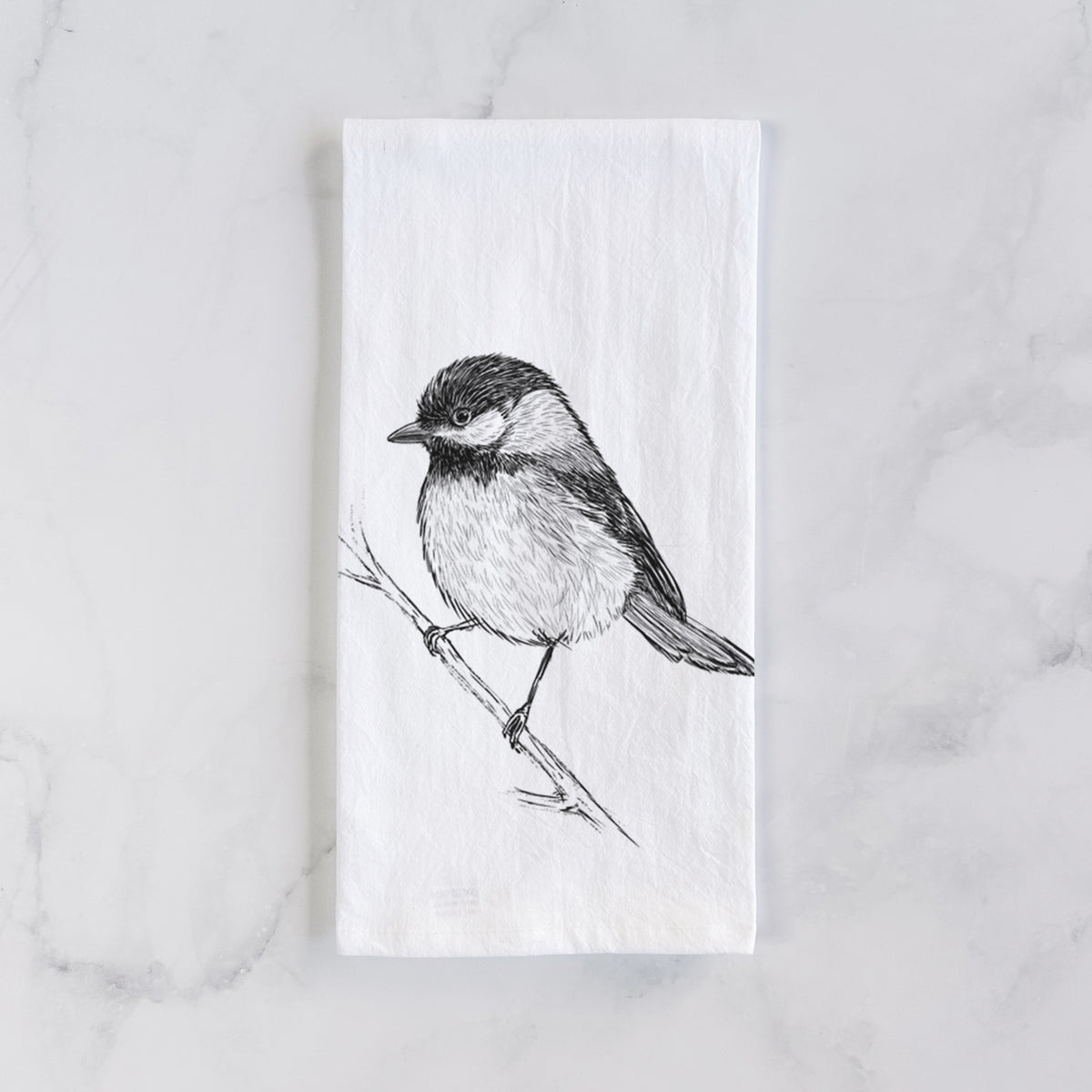 White cotton tea towel laid flat on marble, printed with a detailed black ink sketch of a black-capped chickadee perched on a branch.