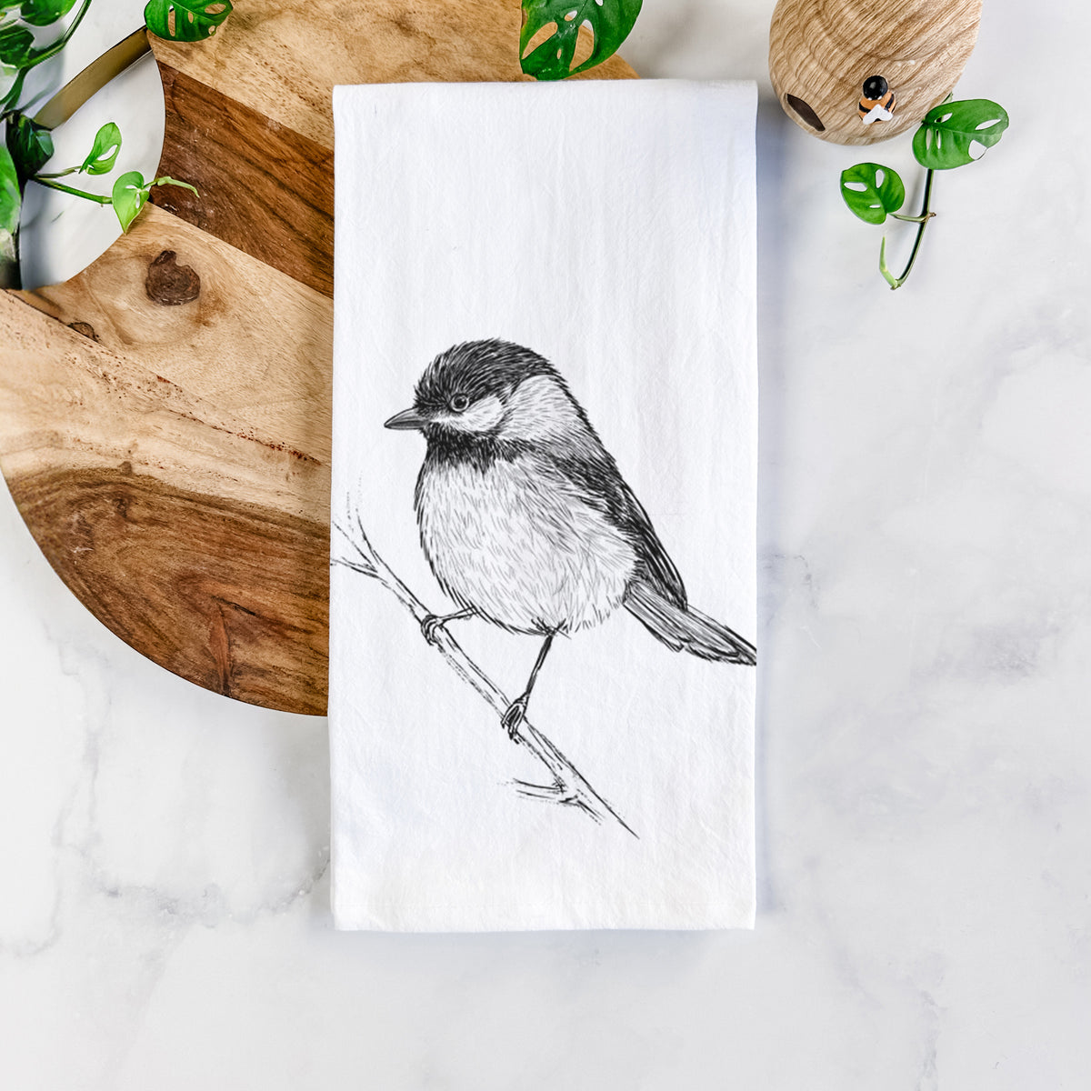 White cotton tea towel printed with a detailed black ink sketch of a black-capped chickadee perched on a branch, on a marble counter beside a round wooden cutting board and greenery.