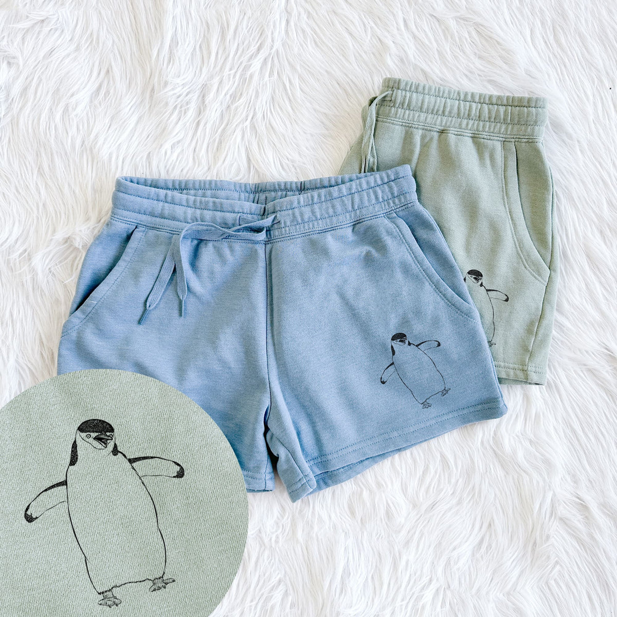 Two pairs of Cali Wave shorts in different colors printed with a black ink illustration of a chinstrap penguin standing with its flippers out, with a close-up of the print.
