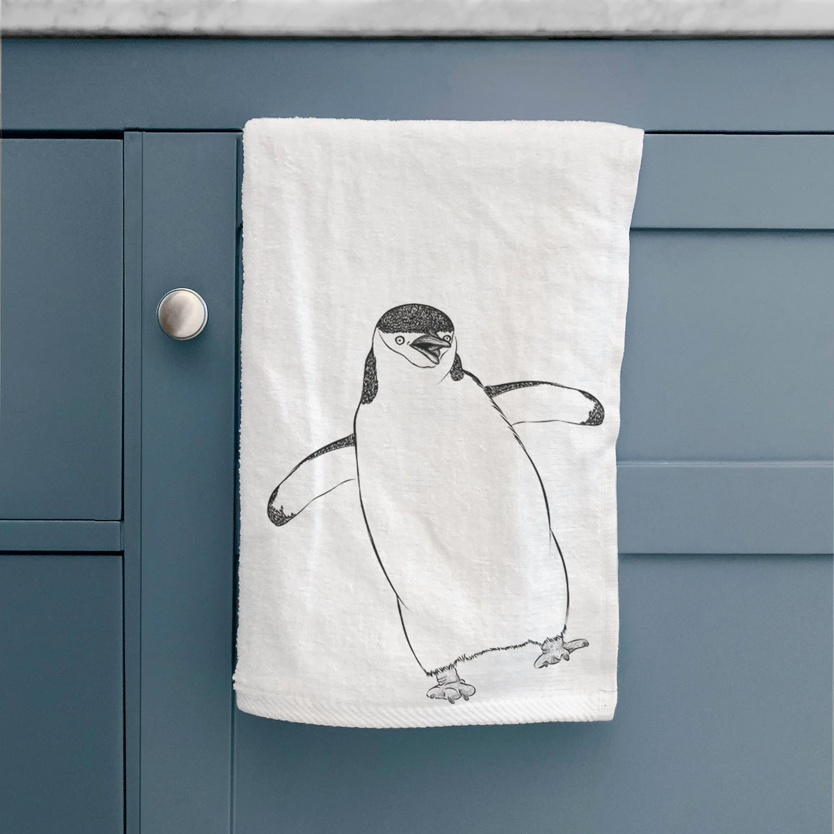 White hand towel printed with a black ink illustration of a chinstrap penguin standing with its flippers out, hanging on a cabinet door.