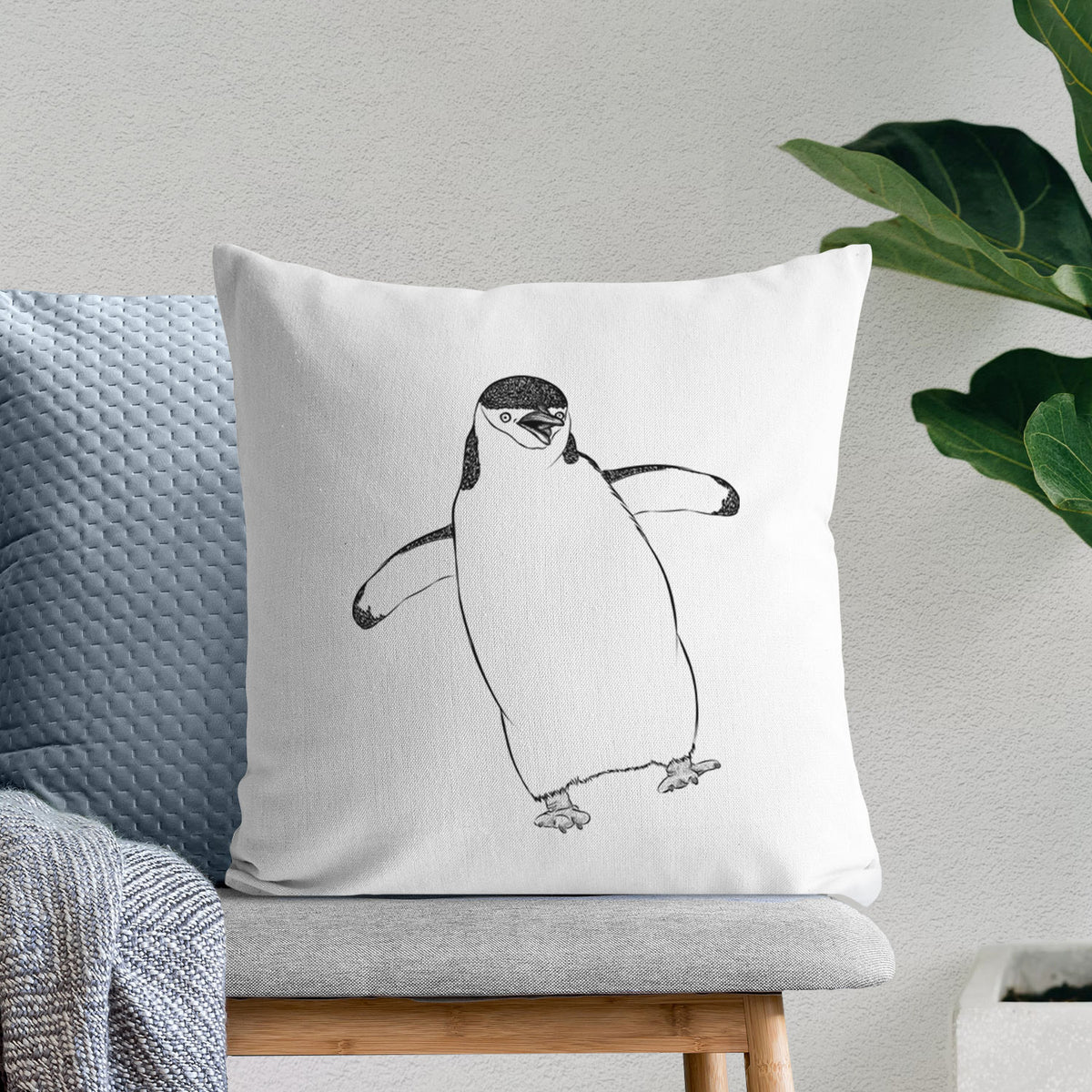 White pillow cover printed with a black ink illustration of a chinstrap penguin standing with its flippers out, on a bench beside plants.