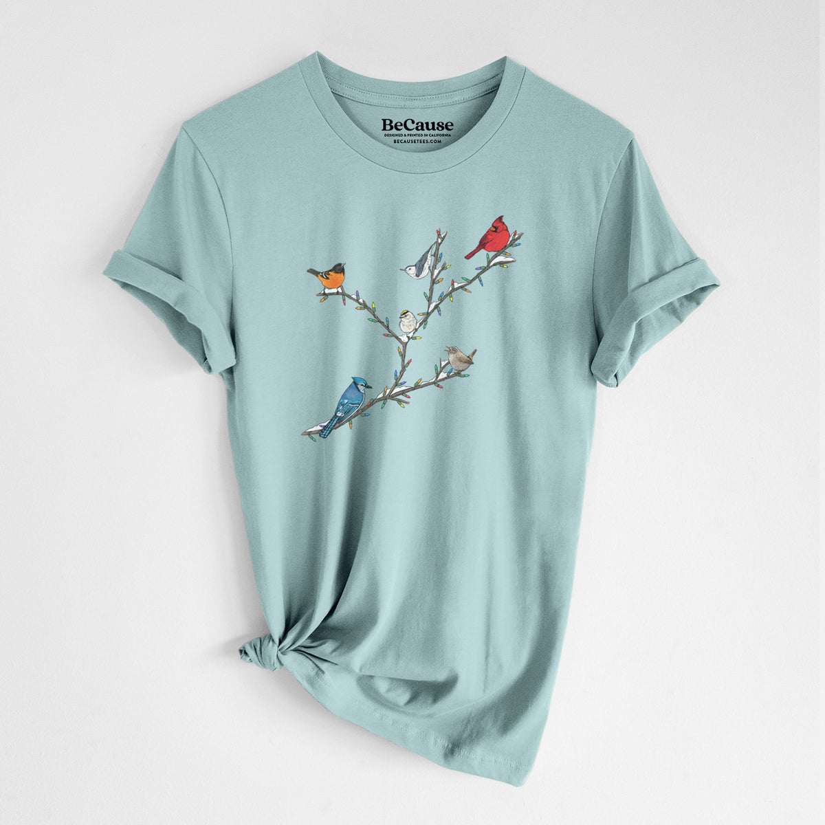 Dusty blue lightweight cotton t-shirt laid flat with the sleeves rolled, printed with a full-color illustration of winter backyard birds, including a cardinal, blue jay and robin, perched on bare branches.