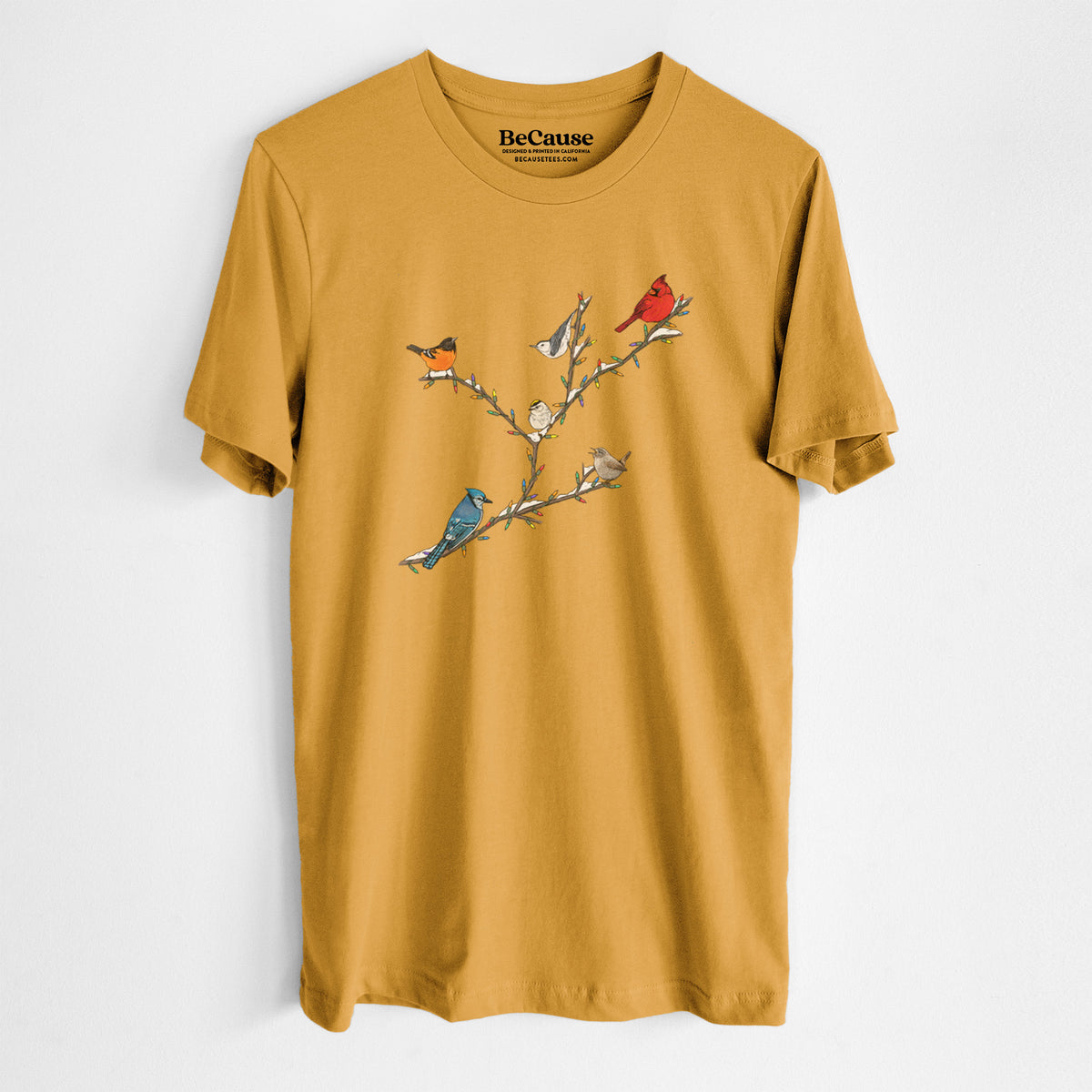 Mustard yellow lightweight cotton t-shirt laid flat, printed with a full-color illustration of winter backyard birds, including a cardinal, blue jay and robin, perched on bare branches.