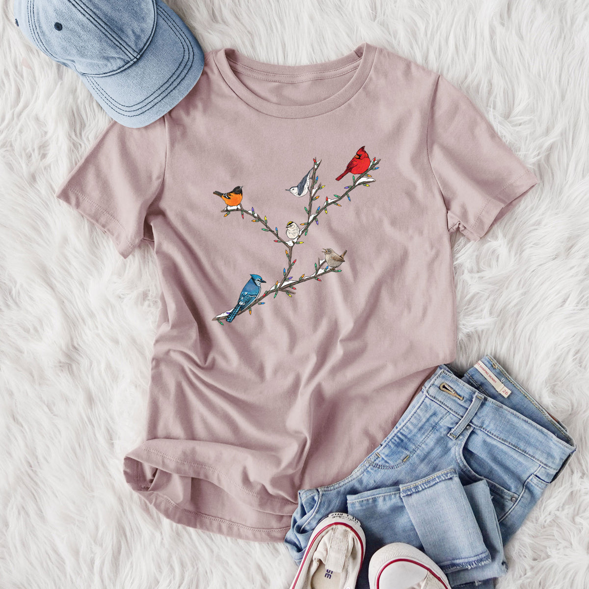 Women's relaxed fit cotton t-shirt printed with a full-color illustration of winter backyard birds, including a cardinal, blue jay and robin, perched on bare branches, styled flat on a white faux fur rug with jeans.