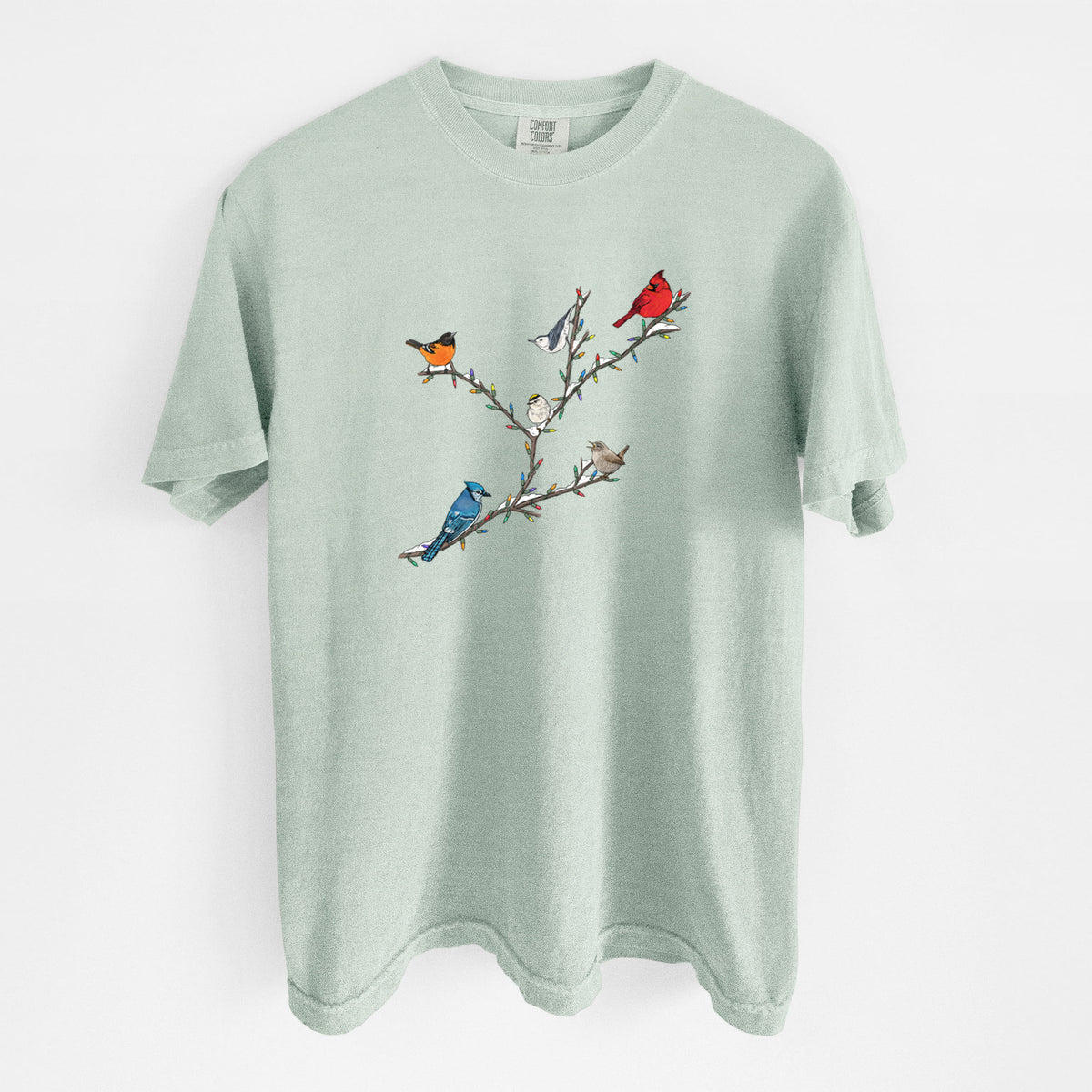 Bay green Comfort Colors t-shirt laid flat, printed with a full-color illustration of winter backyard birds, including a cardinal, blue jay and robin, perched on bare branches.