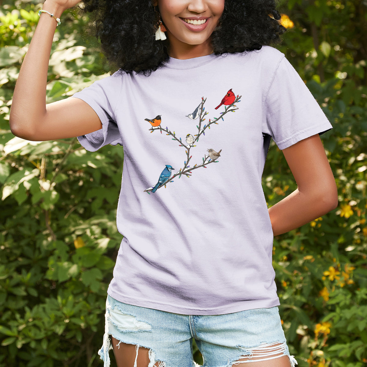 Woman wearing a Comfort Colors t-shirt printed with a full-color illustration of winter backyard birds, including a cardinal, blue jay and robin, perched on bare branches, outdoors.