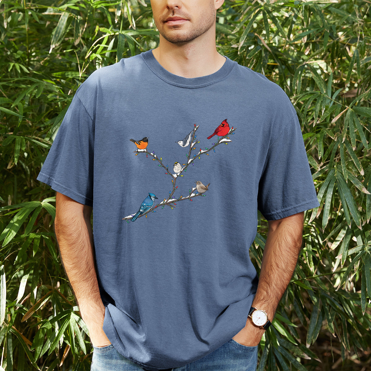 Man wearing a Comfort Colors t-shirt printed with a full-color illustration of winter backyard birds, including a cardinal, blue jay and robin, perched on bare branches, outdoors.