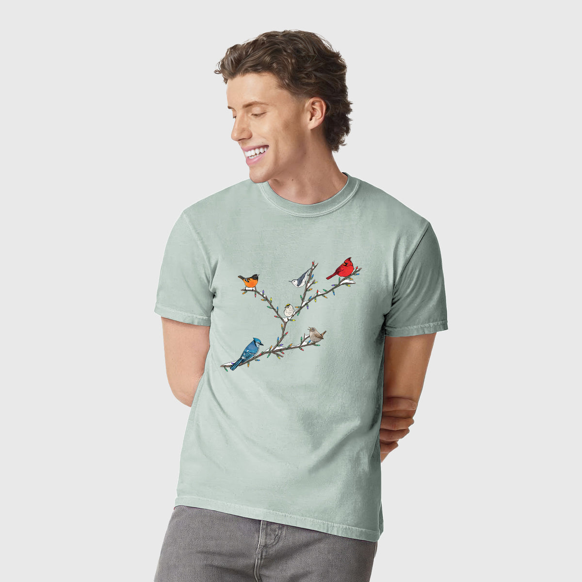 Man wearing a Comfort Colors t-shirt printed with a full-color illustration of winter backyard birds, including a cardinal, blue jay and robin, perched on bare branches.