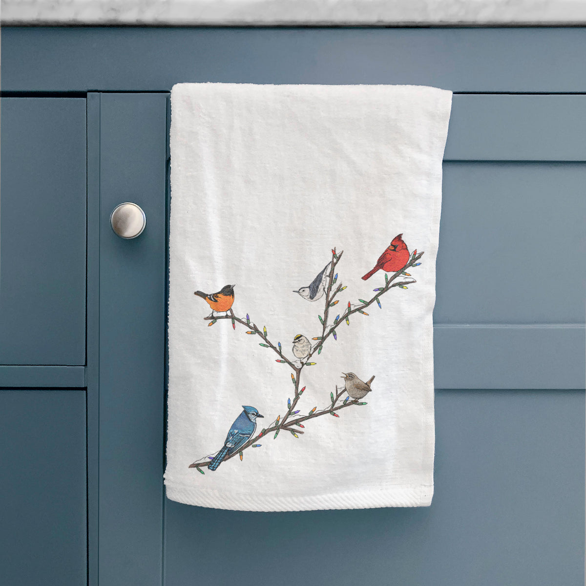 White hand towel printed with a full-color illustration of winter backyard birds, including a cardinal, blue jay and robin, perched on bare branches, hanging on a cabinet door.