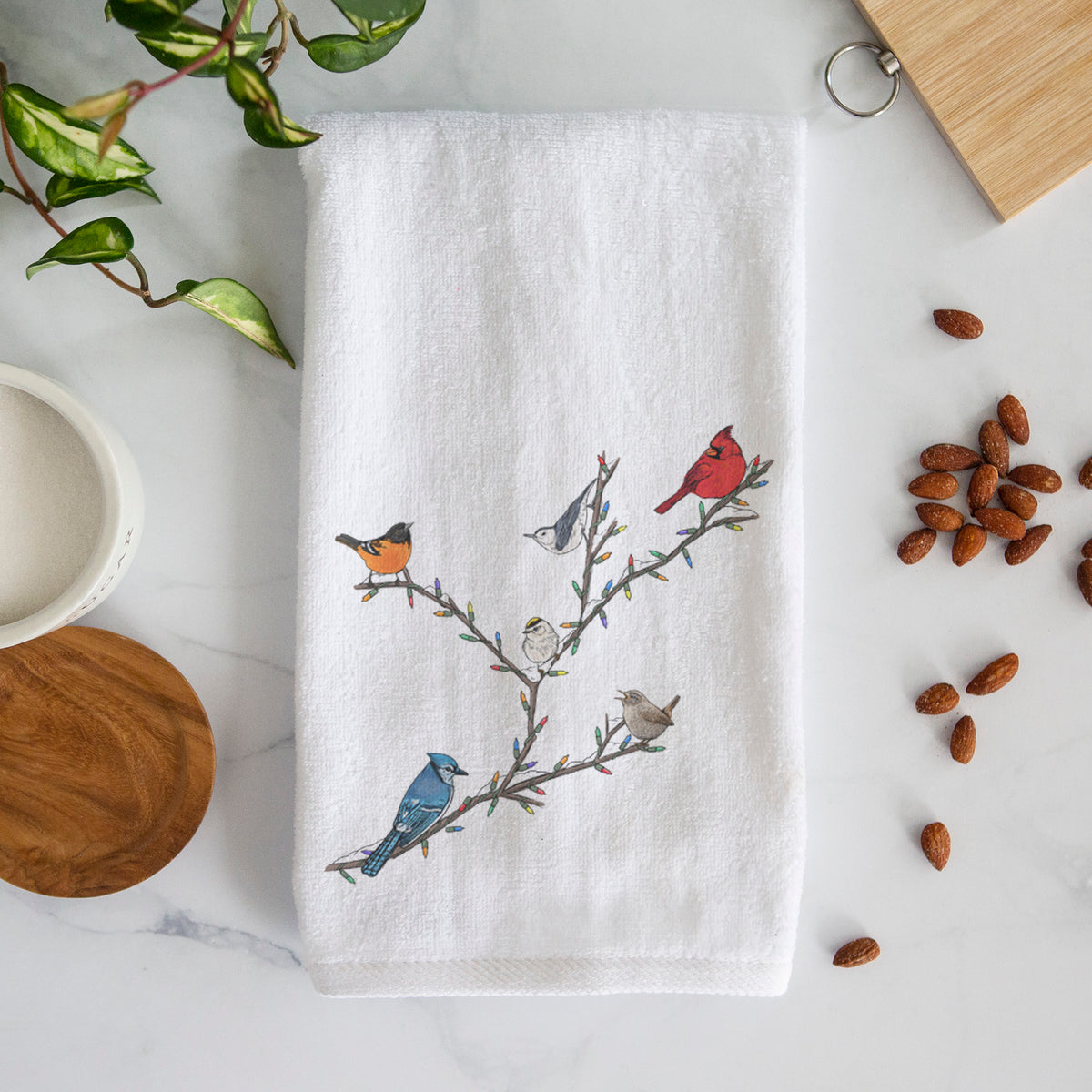 White hand towel printed with a full-color illustration of winter backyard birds, including a cardinal, blue jay and robin, perched on bare branches.