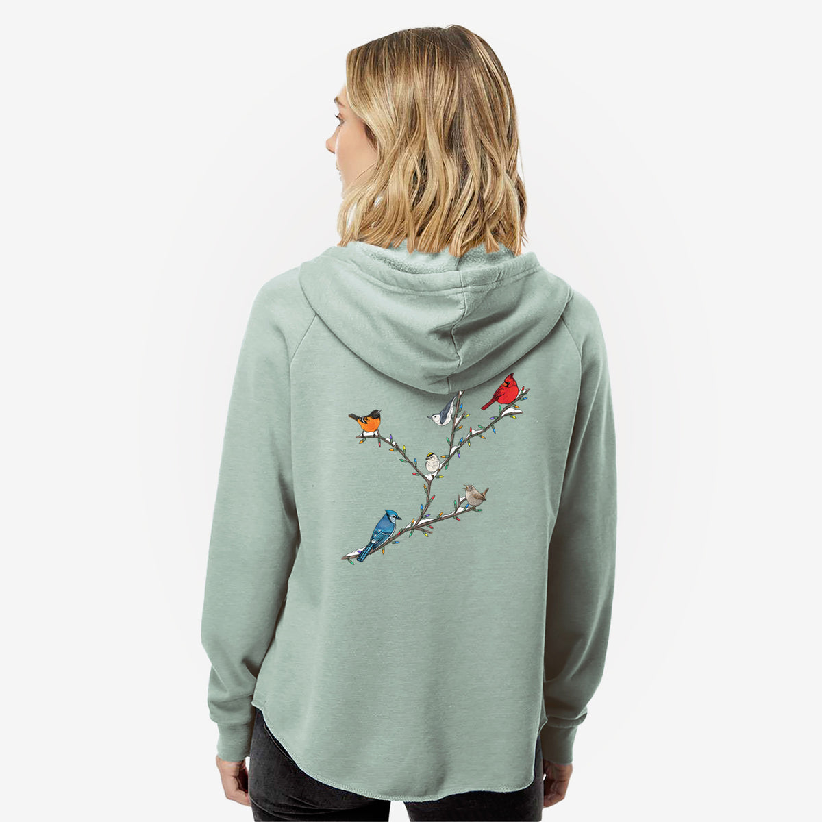 Woman wearing a sage green women's Cali Wave zip-up sweatshirt printed with a full-color illustration of winter backyard birds, including a cardinal, blue jay and robin, perched on bare branches, seen from behind.