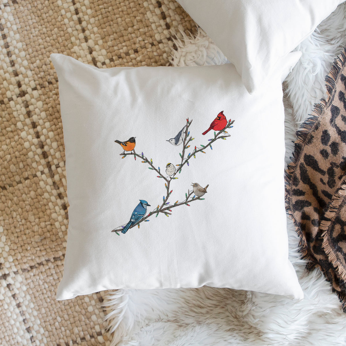 White pillow cover printed with a full-color illustration of winter backyard birds, including a cardinal, blue jay and robin, perched on bare branches, on a woven chair.