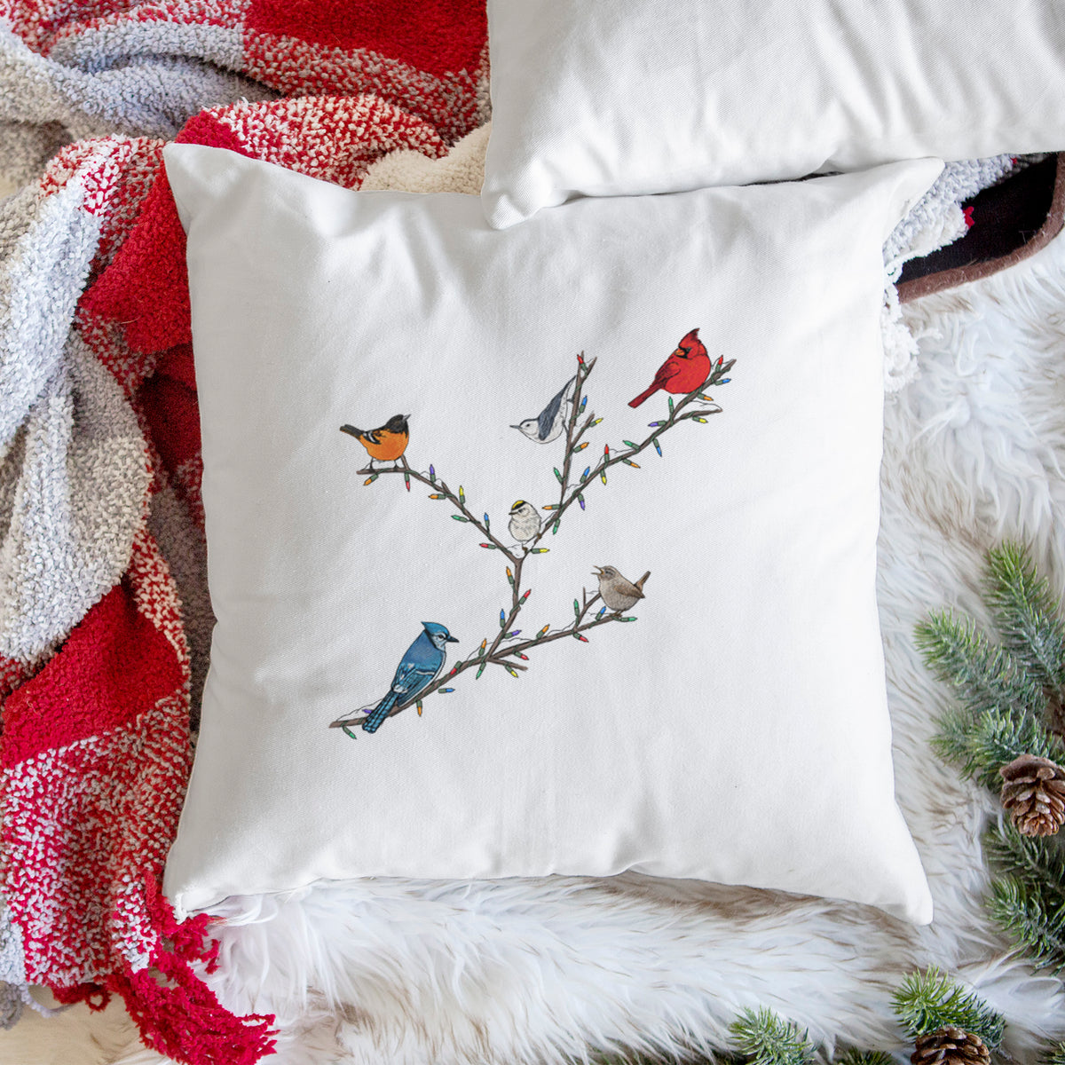 White pillow cover printed with a full-color illustration of winter backyard birds, including a cardinal, blue jay and robin, perched on bare branches, on a woven chair.