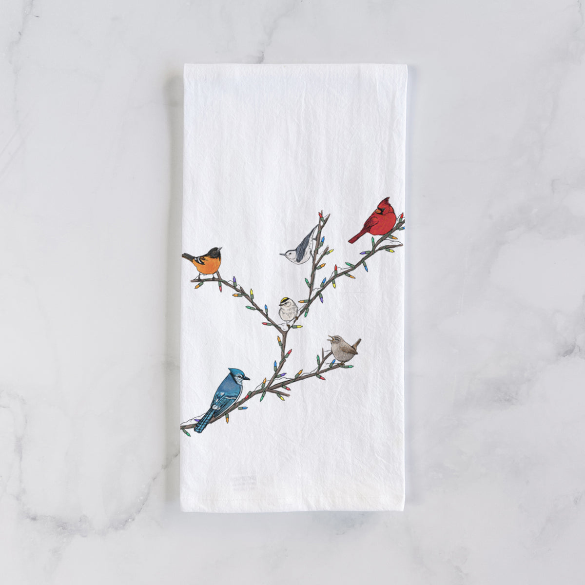 White cotton tea towel laid flat on marble, printed with a full-color illustration of winter backyard birds, including a cardinal, blue jay and robin, perched on bare branches.