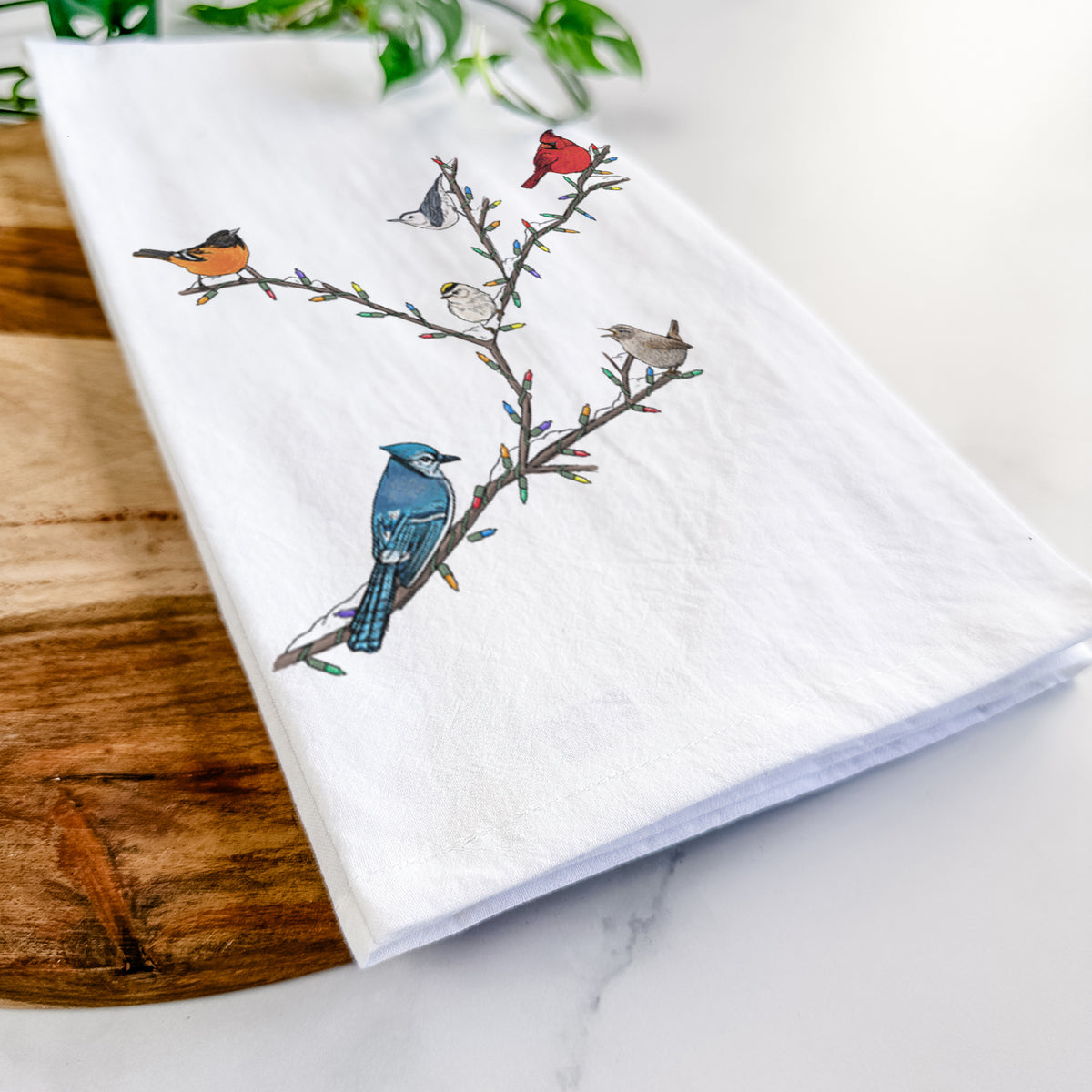 Close-up of a folded white tea towel on a wooden board, showing a full-color illustration of winter backyard birds, including a cardinal, blue jay and robin, perched on bare branches.