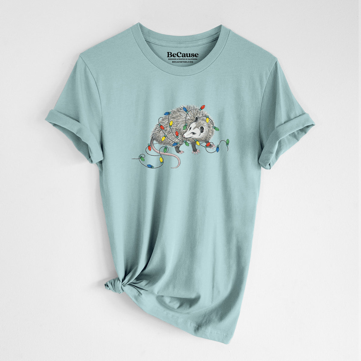Dusty blue lightweight cotton t-shirt laid flat with the sleeves rolled, printed with a black ink opossum tangled in a string of colorful holiday lights.