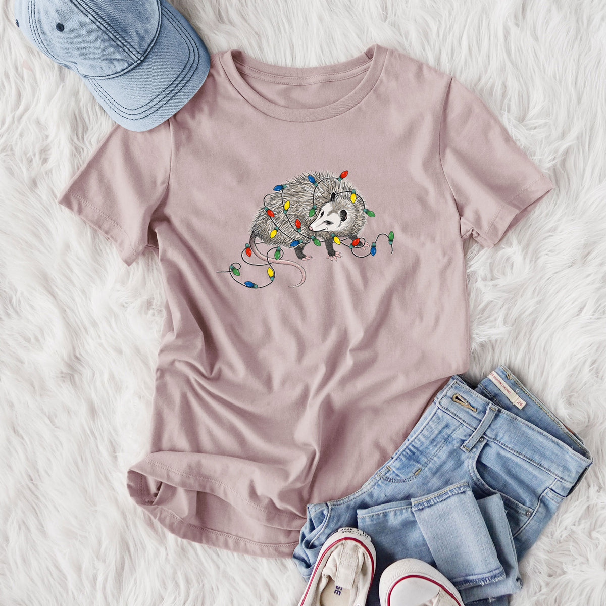 Women's relaxed fit cotton t-shirt printed with a black ink opossum tangled in a string of colorful holiday lights, styled flat on a white faux fur rug with jeans.