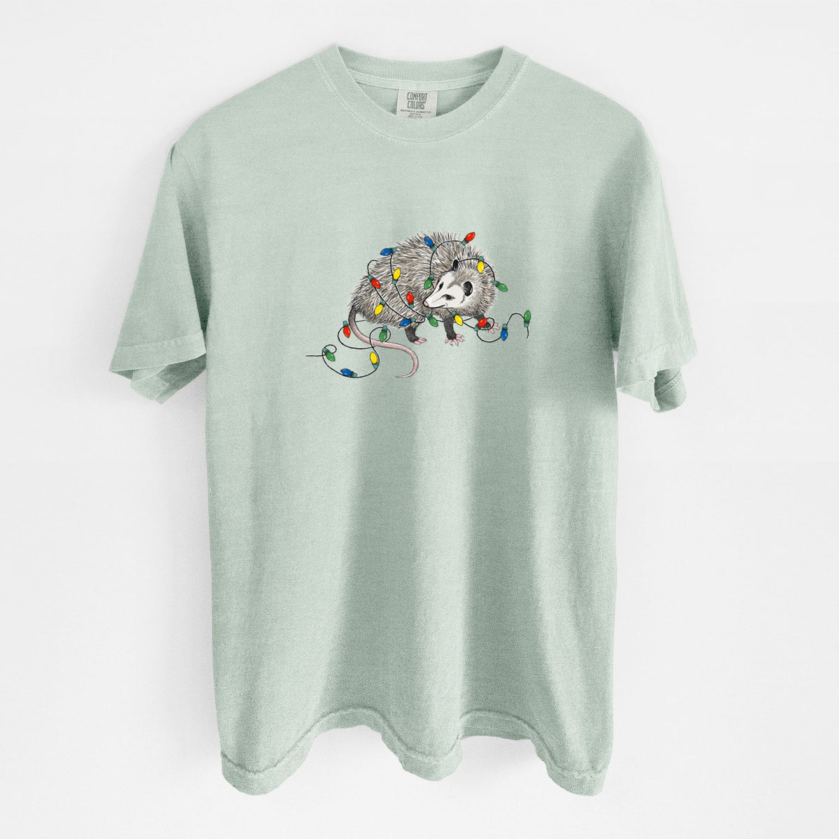 Bay green Comfort Colors t-shirt laid flat, printed with a black ink opossum tangled in a string of colorful holiday lights.