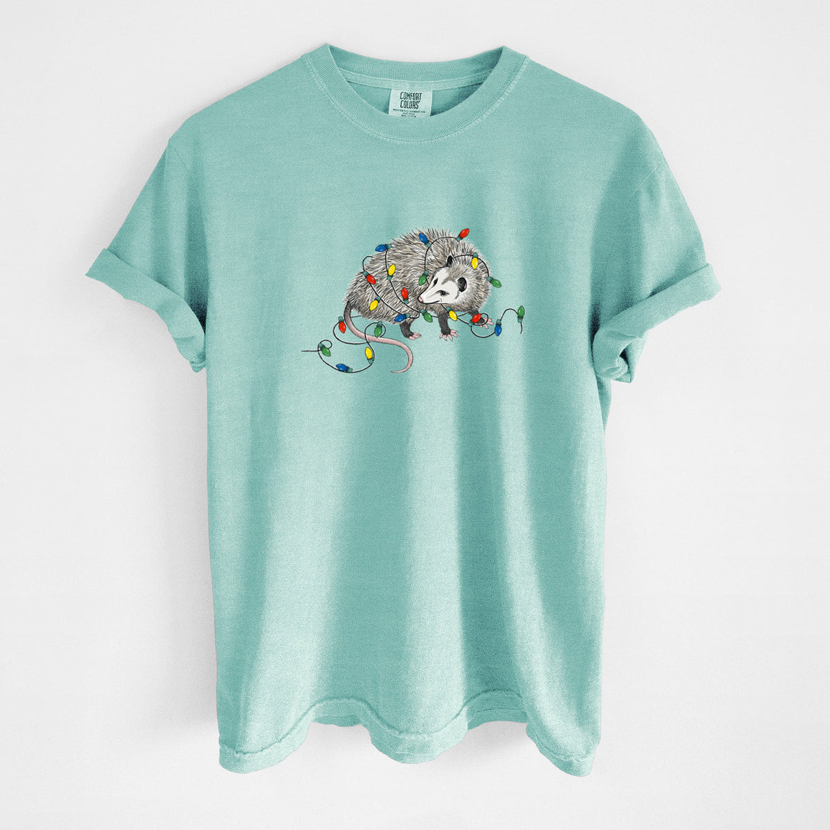 Chalky mint Comfort Colors t-shirt laid flat, printed with a black ink opossum tangled in a string of colorful holiday lights.