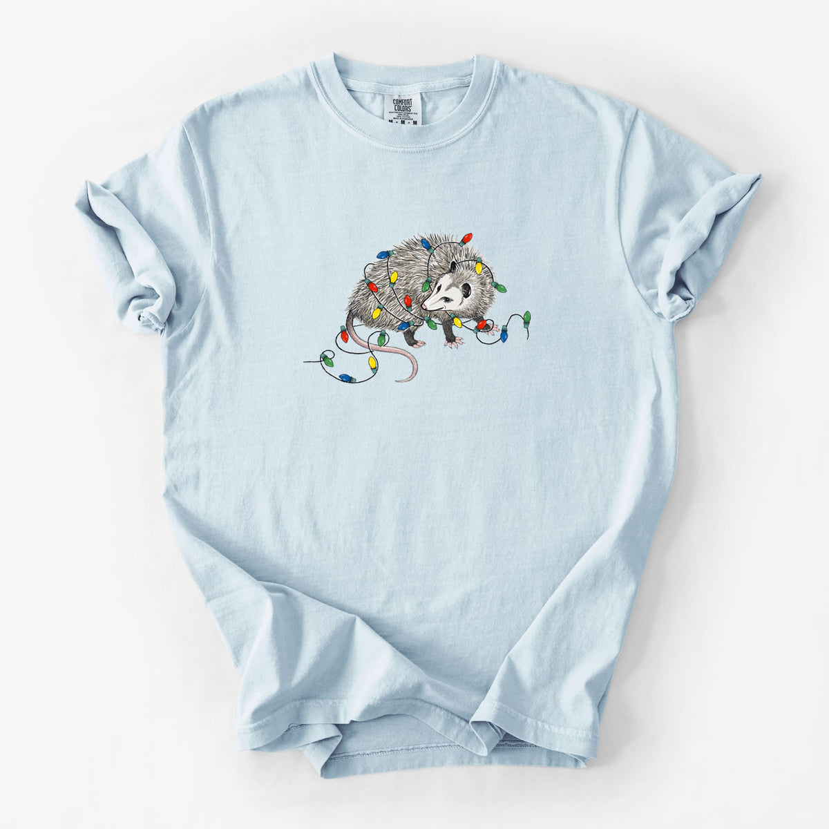 Chambray blue Comfort Colors t-shirt laid flat, printed with a black ink opossum tangled in a string of colorful holiday lights.