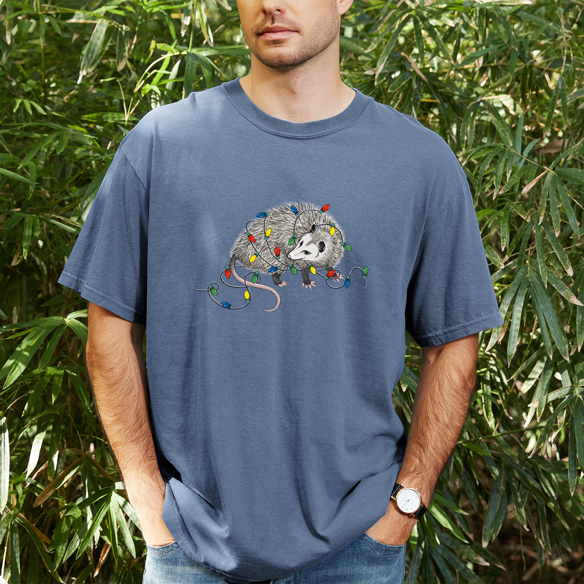 Man wearing a Comfort Colors t-shirt printed with a black ink opossum tangled in a string of colorful holiday lights, outdoors.