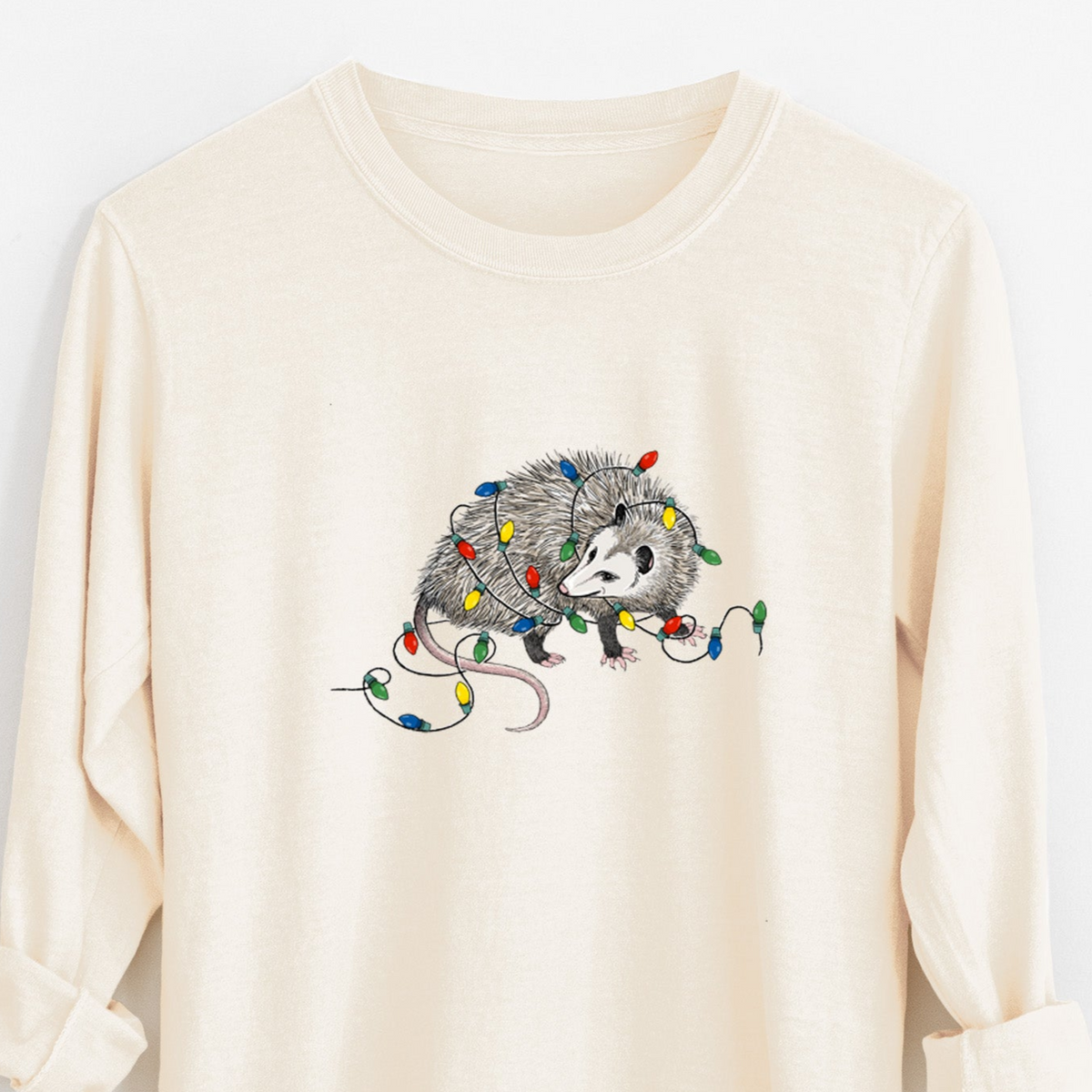 Ivory Comfort Colors long sleeve shirt laid flat with the cuffs rolled, printed with a black ink opossum tangled in a string of colorful holiday lights.
