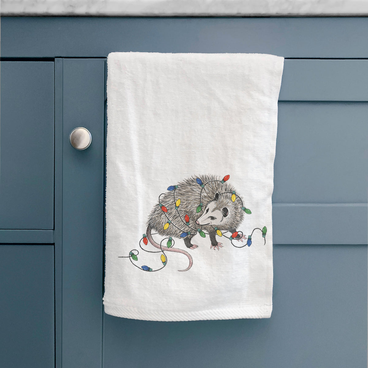 White hand towel printed with a black ink opossum tangled in a string of colorful holiday lights, hanging on a cabinet door.