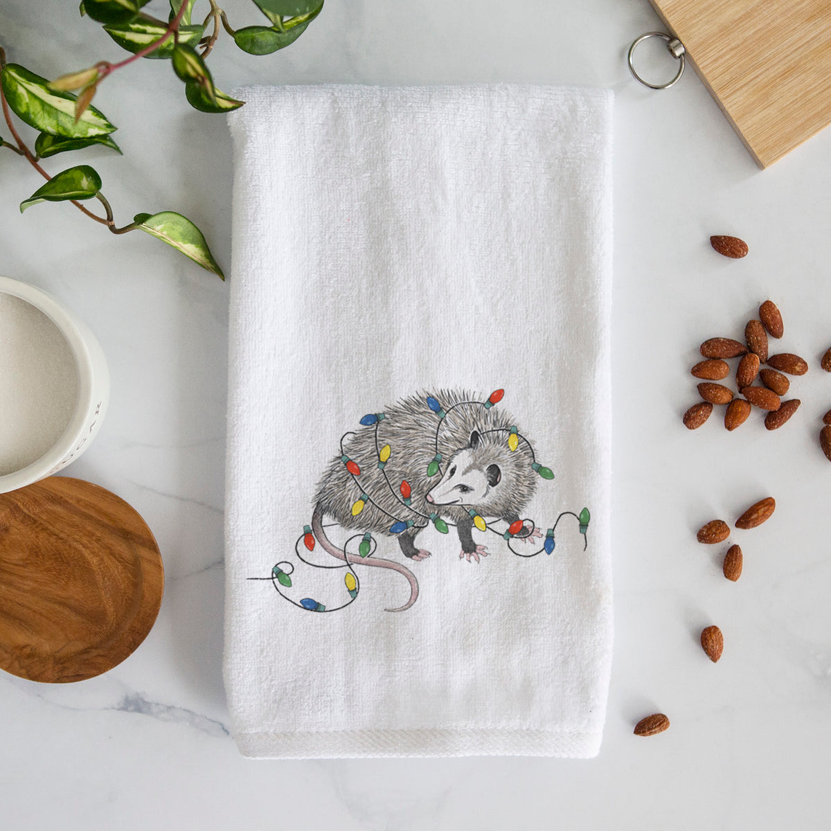 White hand towel printed with a black ink opossum tangled in a string of colorful holiday lights.