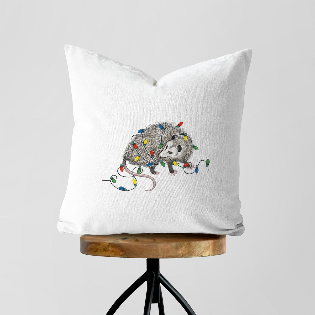 White pillow cover printed with a black ink opossum tangled in a string of colorful holiday lights, on a wooden stool.