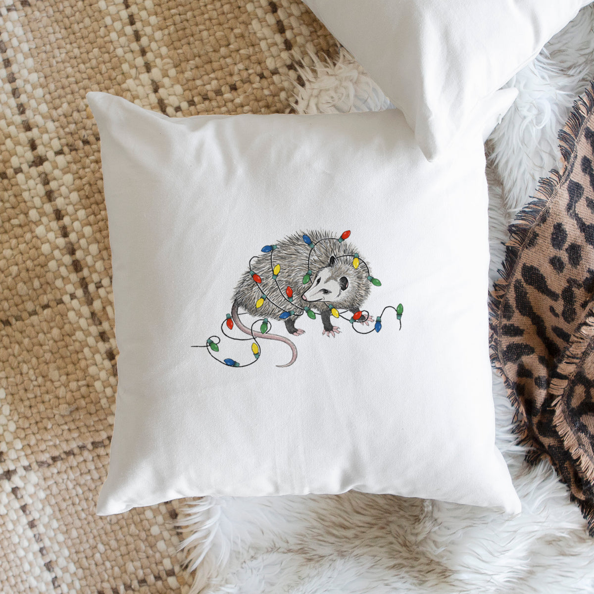 White pillow cover printed with a black ink opossum tangled in a string of colorful holiday lights, on a woven chair.