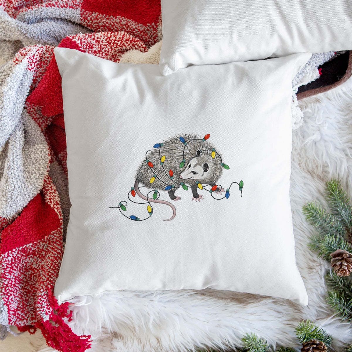 White pillow cover printed with a black ink opossum tangled in a string of colorful holiday lights, on a woven chair.