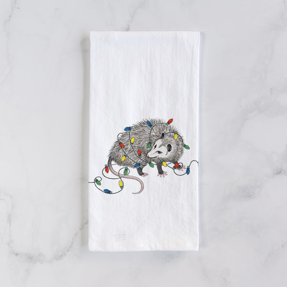 White cotton tea towel laid flat on marble, printed with a black ink opossum tangled in a string of colorful holiday lights.