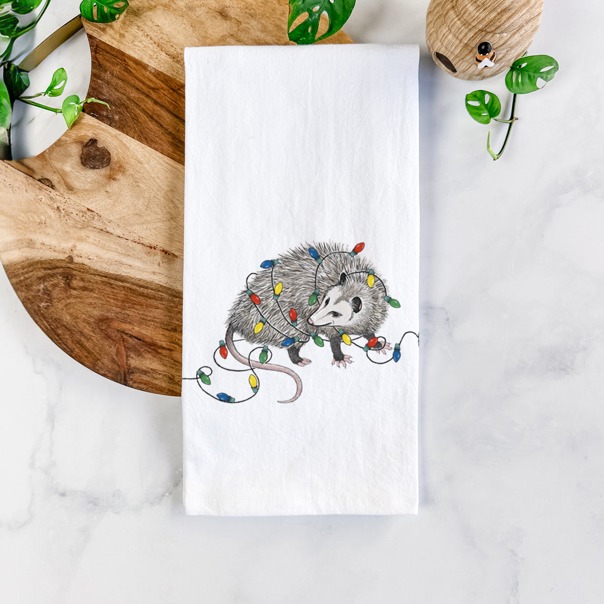 White cotton tea towel printed with a black ink opossum tangled in a string of colorful holiday lights, on a marble counter beside a round wooden cutting board and greenery.