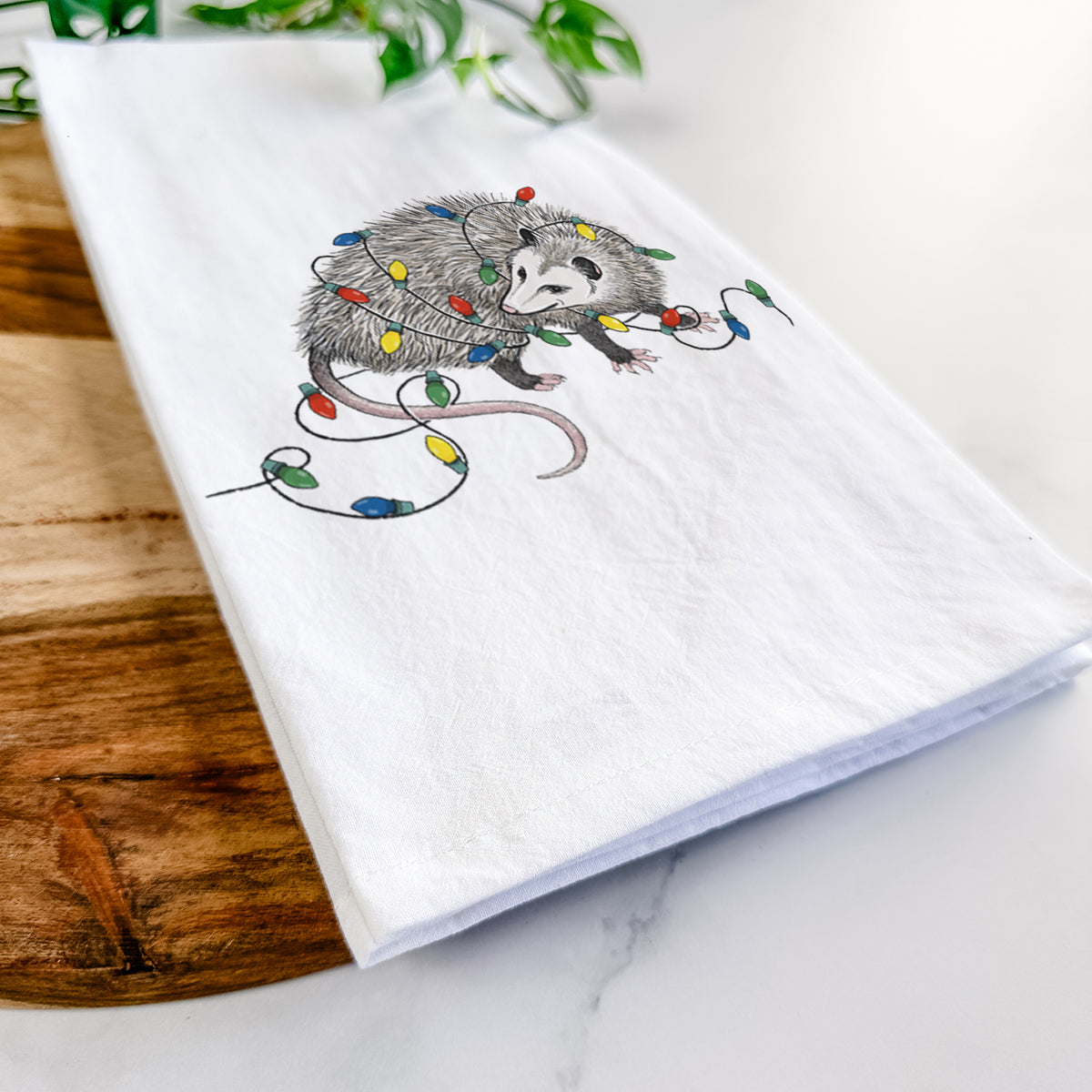 Close-up of a folded white tea towel on a wooden board, showing a black ink opossum tangled in a string of colorful holiday lights.