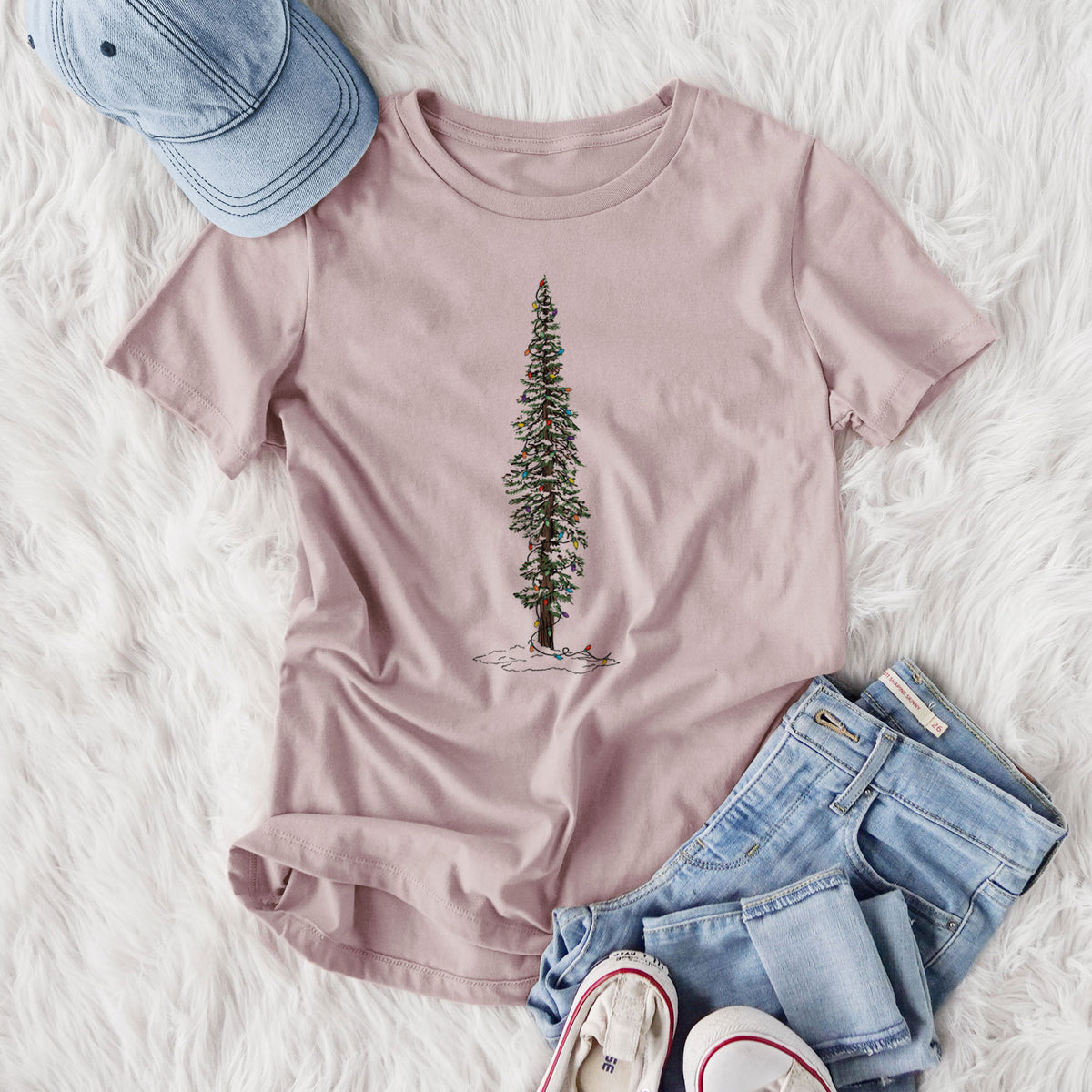 Women's relaxed fit cotton t-shirt printed with a full-color illustration of a tall redwood tree dusted with snow and strung with holiday lights, styled flat on a white faux fur rug with jeans.