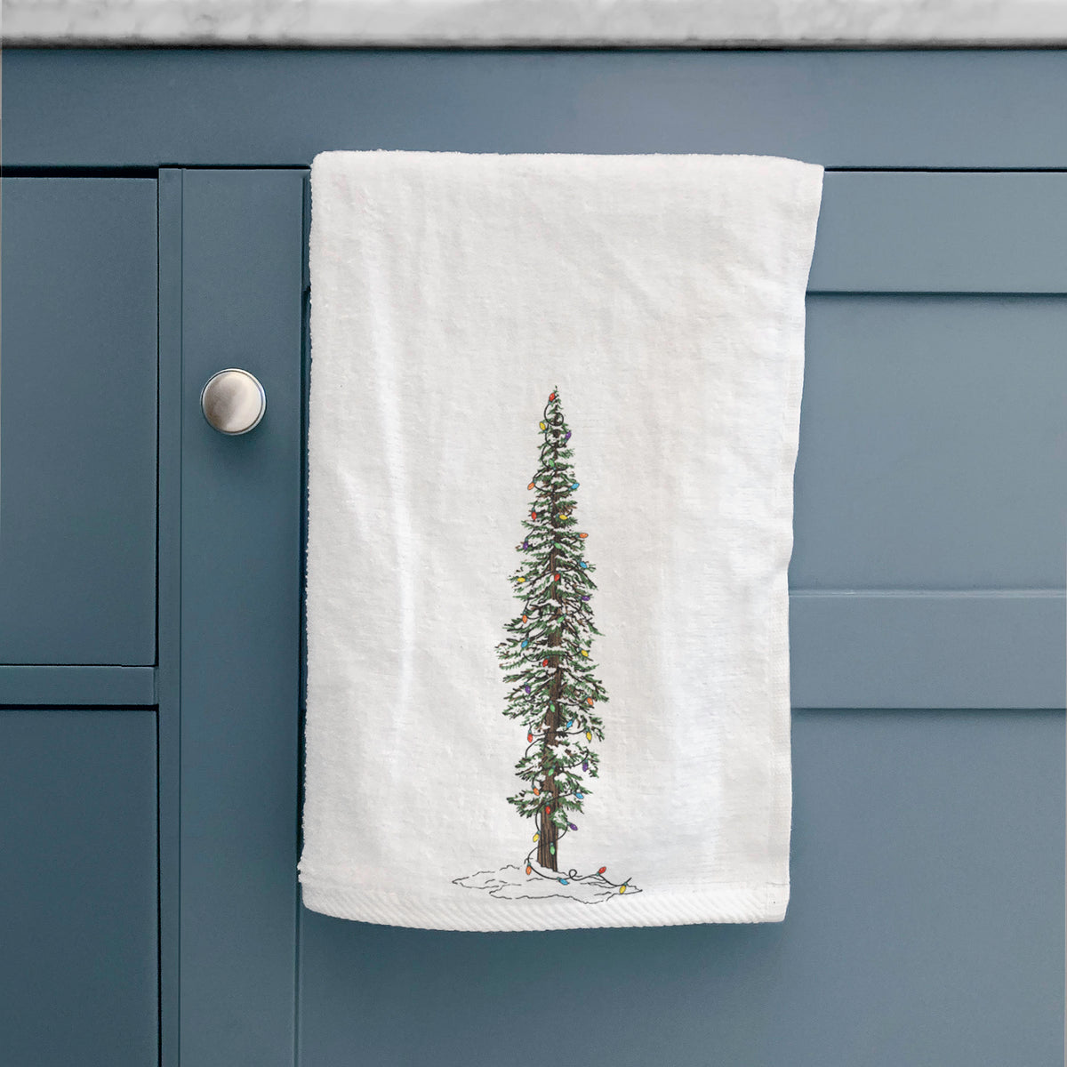 White hand towel printed with a full-color illustration of a tall redwood tree dusted with snow and strung with holiday lights, hanging on a cabinet door.