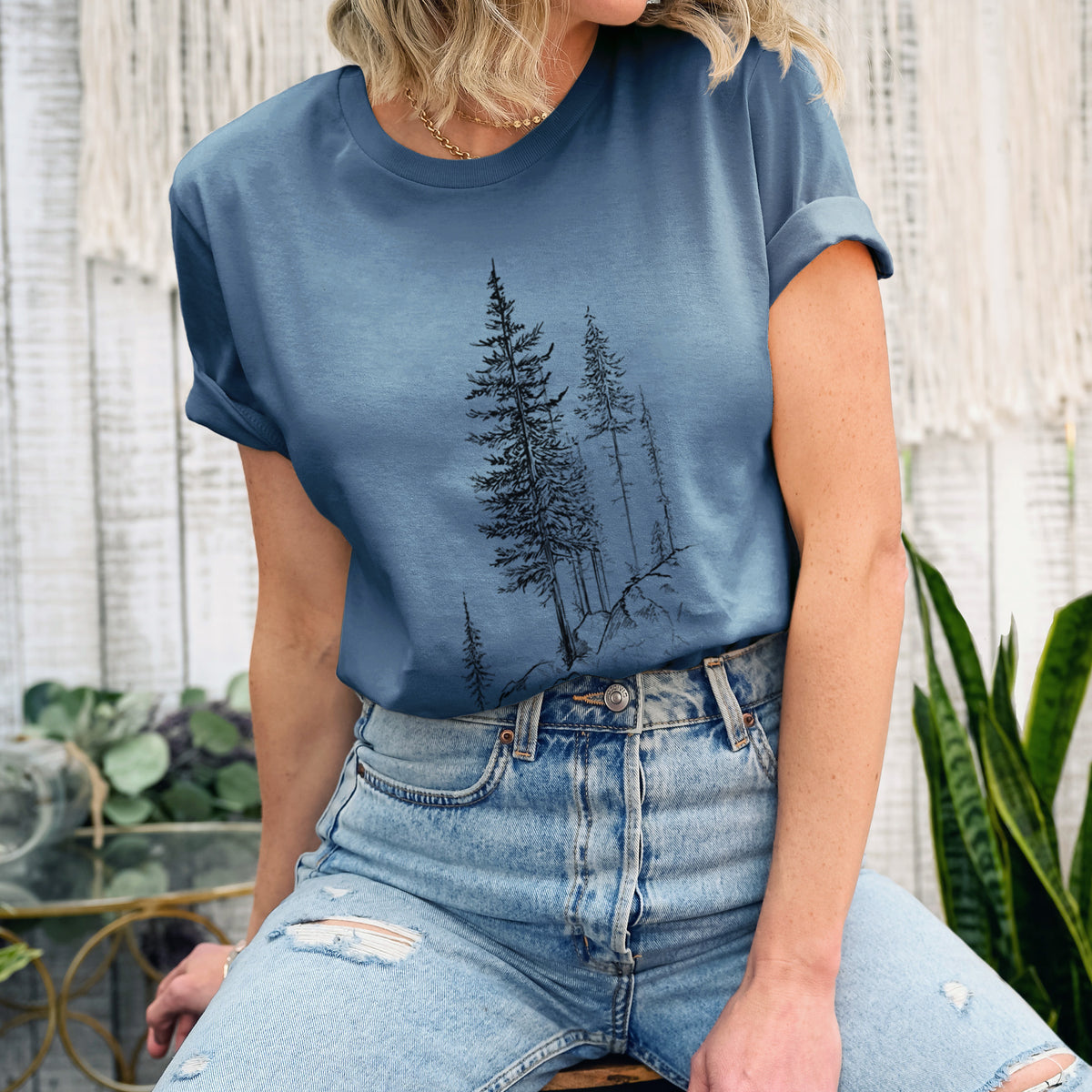 Woman wearing a steel blue lightweight cotton t-shirt printed with a black ink illustration of tall pine trees growing from a rocky cliff.