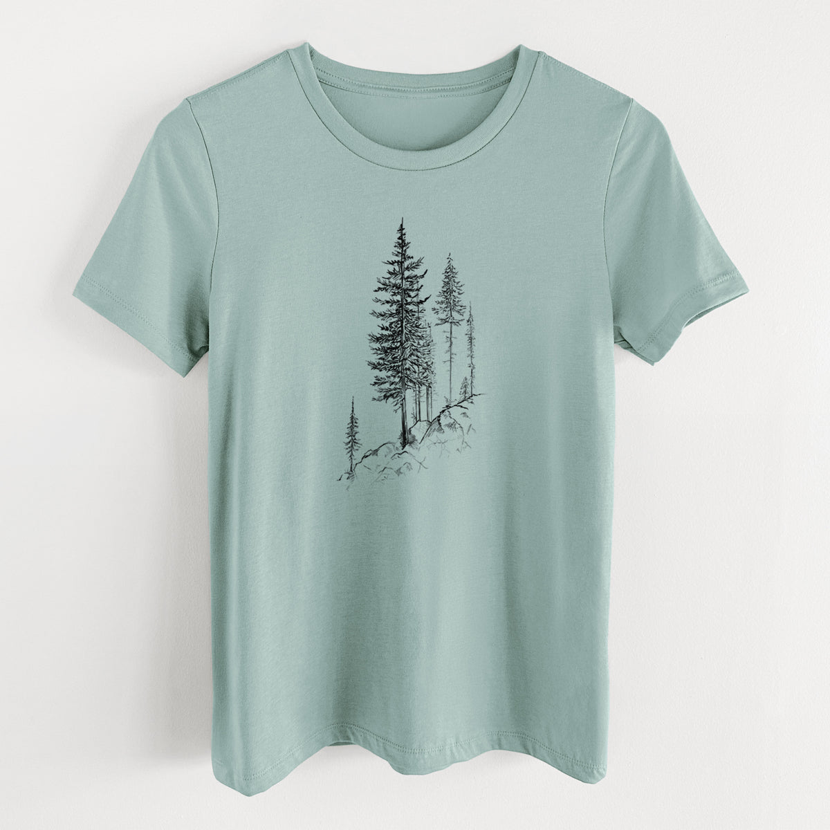 Dusty blue women's relaxed fit cotton t-shirt laid flat, printed with a black ink illustration of tall pine trees growing from a rocky cliff.