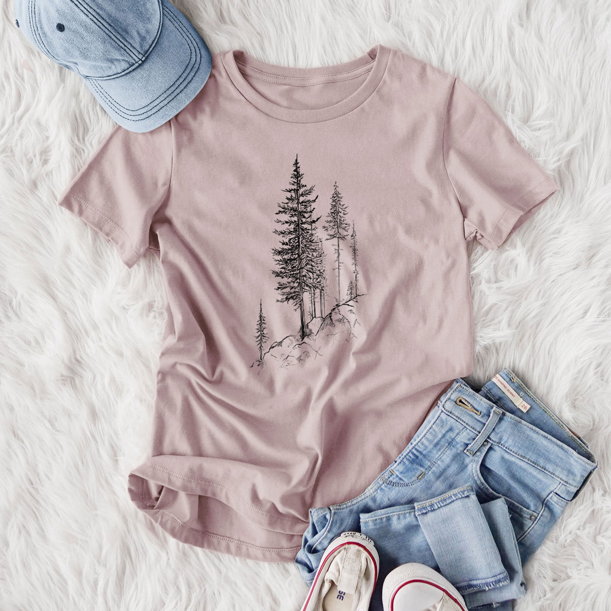 Women's relaxed fit cotton t-shirt printed with a black ink illustration of tall pine trees growing from a rocky cliff, styled flat on a white faux fur rug with jeans.