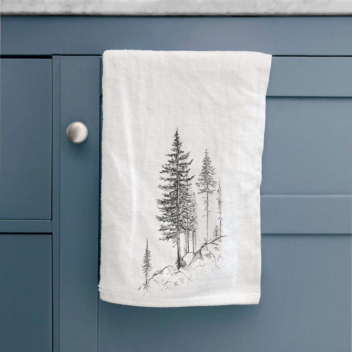 White hand towel printed with a black ink illustration of tall pine trees growing from a rocky cliff, hanging on a cabinet door.