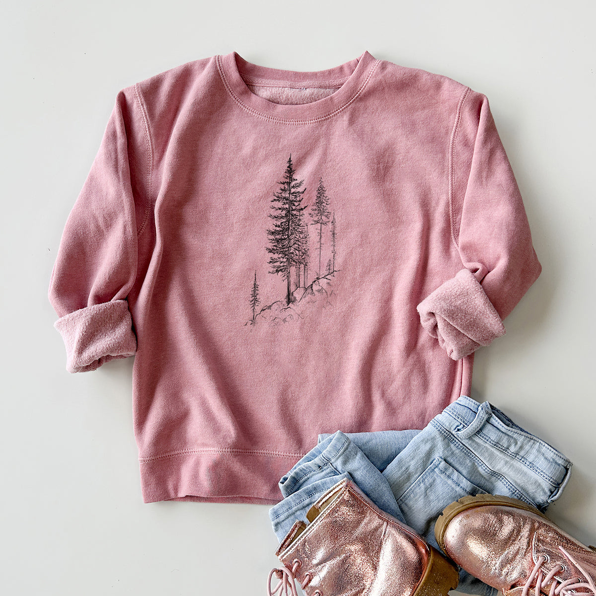 Youth crewneck sweatshirt printed with a black ink illustration of tall pine trees growing from a rocky cliff, styled flat with jeans and boots.