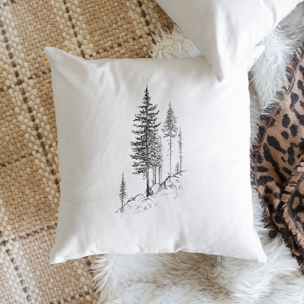 White pillow cover printed with a black ink illustration of tall pine trees growing from a rocky cliff, on a woven chair.