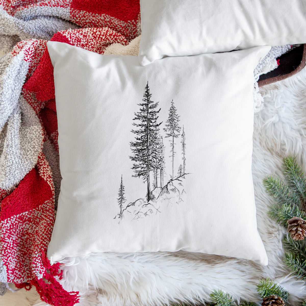White pillow cover printed with a black ink illustration of tall pine trees growing from a rocky cliff, on a woven chair.