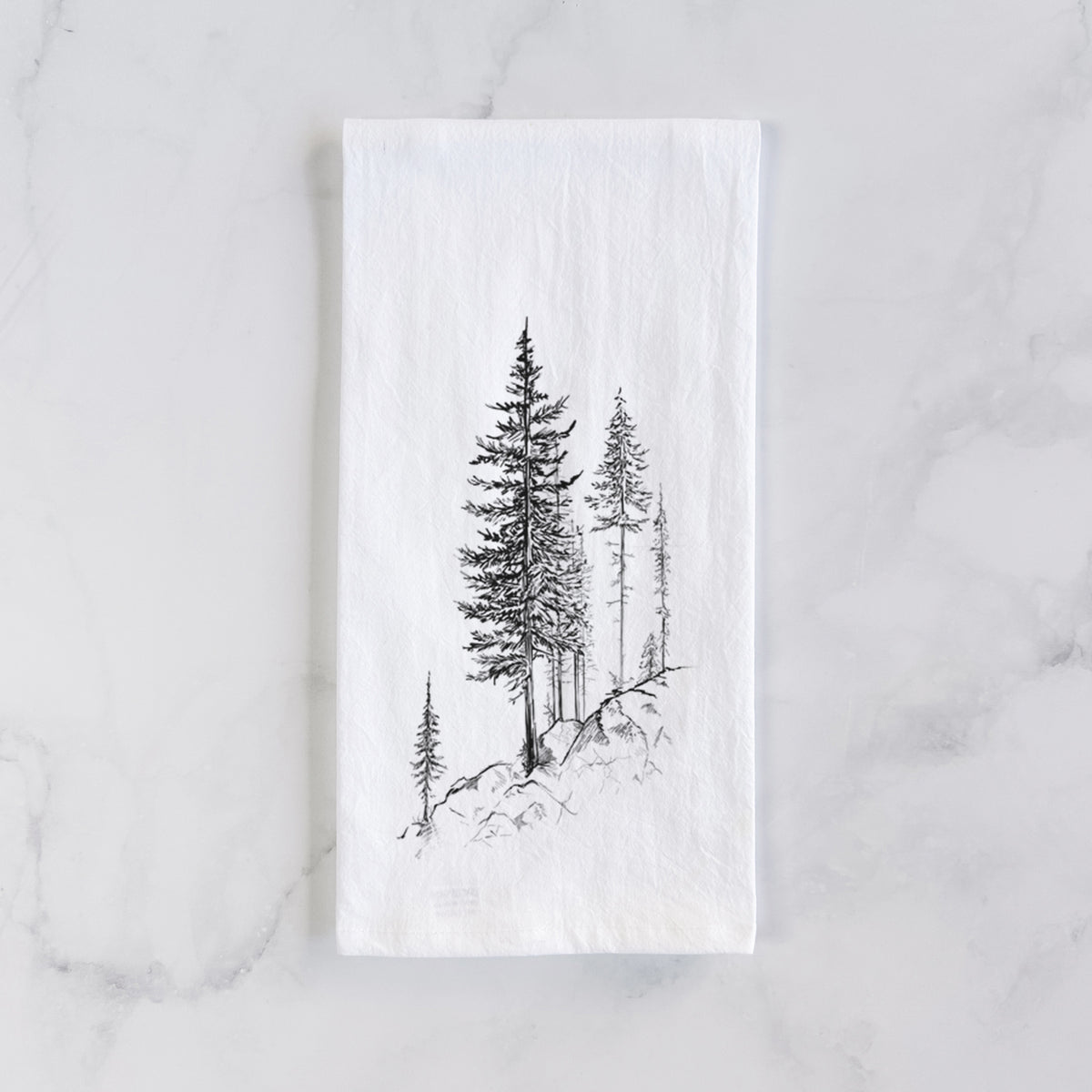 White cotton tea towel laid flat on marble, printed with a black ink illustration of tall pine trees growing from a rocky cliff.
