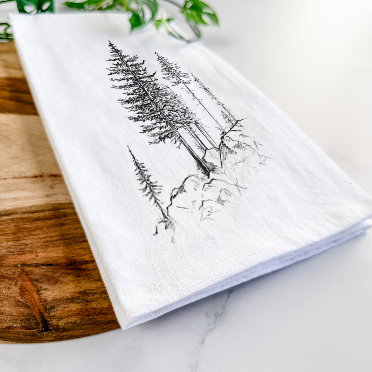 Close-up of a folded white tea towel on a wooden board, showing a black ink illustration of tall pine trees growing from a rocky cliff.
