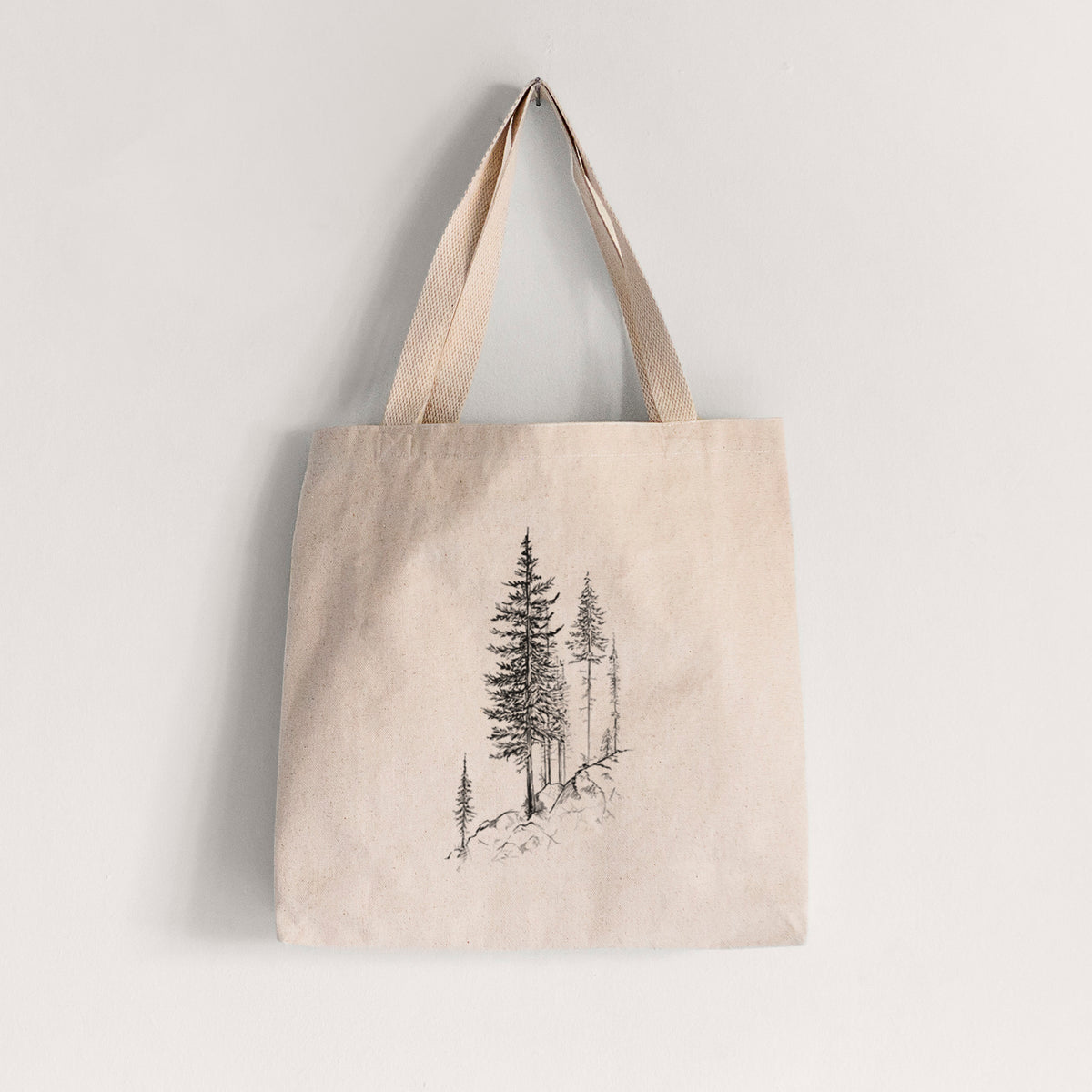 Natural canvas tote bag printed with a black ink illustration of tall pine trees growing from a rocky cliff.